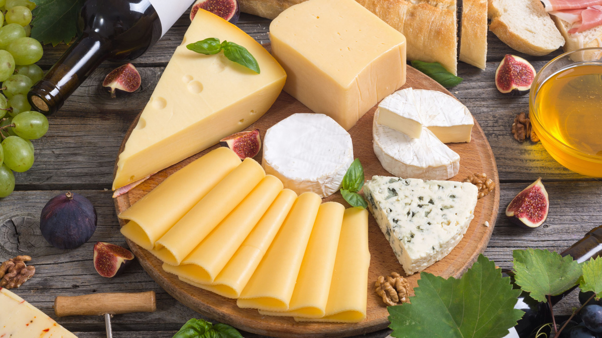KI generiert: Das Bild zeigt ein hölzernes Brett mit einer Auswahl an verschiedenen Käsesorten, Trauben, Nüssen und Baguettescheiben. Daneben stehen zwei Weingläser, eines davon mit Rotwein gefüllt.