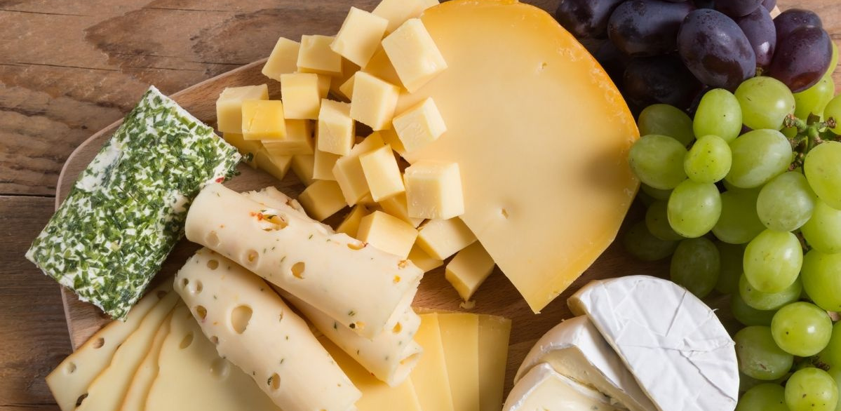 KI generiert: Das Bild zeigt ein hölzernes Brett mit einer Auswahl an verschiedenen Käsesorten, Trauben, Nüssen und Baguettescheiben. Daneben stehen zwei Weingläser, eines davon mit Rotwein gefüllt.