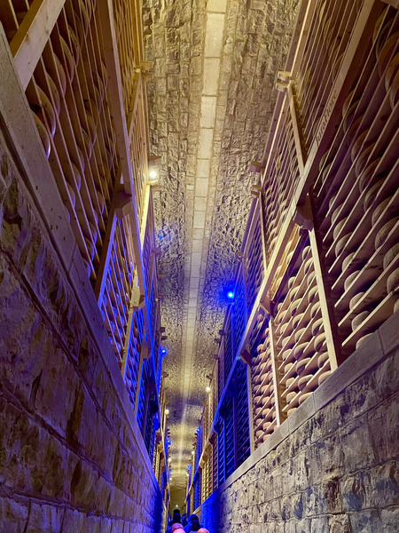 KI generiert: Ein schmaler Weg in einem Weinkeller mit vielen Weinregalen und gedämpfter Beleuchtung.