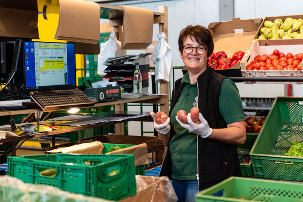 Das Bild zeigt eine Frau, die in einem Geschäft vor einem Regal mit Bio-Produkten steht. Sie trägt ein blaues Oberteil und hat blonde Haare. Sie lächelt freundlich in die Kamera. Im Hintergrund sind Regale mit verschiedenen Produkten zu sehen, die auf eine natürliche und nachhaltige Präsentation hindeuten.