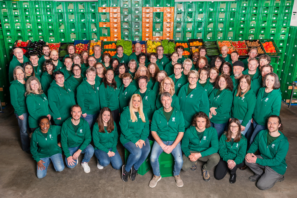 KI generiert: Eine große Gruppe in grünen Hoodies posiert vor Gemüsekisten; die Kisten formen die Zahl "30".