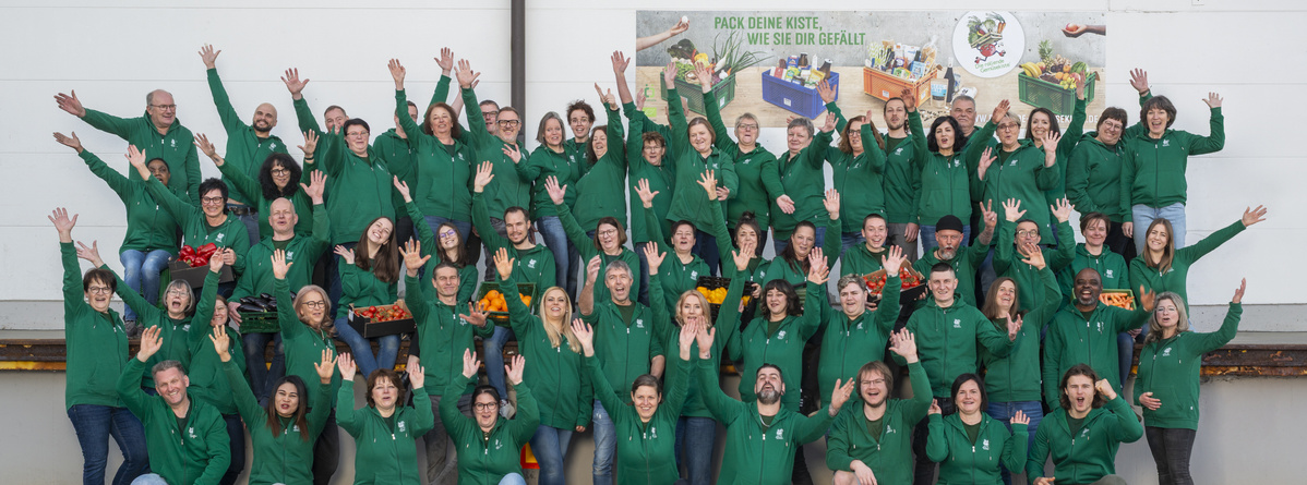 KI generiert: Eine Gruppe von Menschen in grünen Hoodies posiert freudig zusammen. Text: "PACK DEINE KISTE, WIE SIE DIR GEFÄLLT."