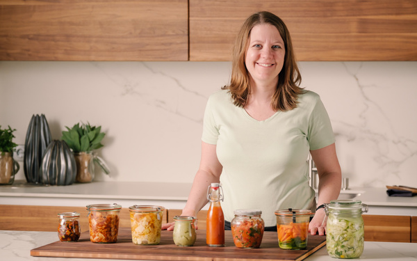 KI generiert: Das Bild zeigt zwei Personen in grüner Arbeitskleidung, die lächelnd eine Kiste mit frischem Obst und Gemüse halten. Im Hintergrund sind weitere Kisten mit Tomaten, Äpfeln und Salat zu sehen, was auf ein Lager oder eine Verkaufsstelle für frische Lebensmittel hinweist.
