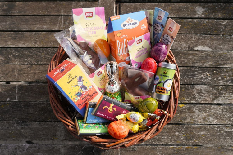 KI generiert: Ein Osterkorb mit Schokolade und Eiern auf Holztisch. Text: "Schoko Milchcreme Eier", "Oster Überraschung", "Gute Laune Mandeln".