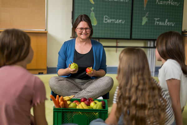 KI generiert: Eine Frau sitzt mit Obst vor einer Tafel, Kinder zuhören. Text: "Birnen, Trauben".