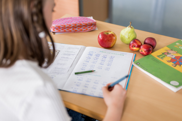 KI generiert: Ein Kind macht Hausaufgaben, mit Schreibheft, Früchten und einem Buch auf dem Tisch.