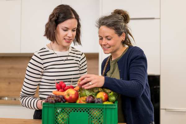 KI generiert: Das Bild zeigt zwei Personen in grüner Arbeitskleidung, die lächelnd eine Kiste mit frischem Obst und Gemüse halten. Im Hintergrund sind weitere Kisten mit Tomaten, Äpfeln und Salat zu sehen, was auf ein Lager oder eine Verkaufsstelle für frische Lebensmittel hinweist.