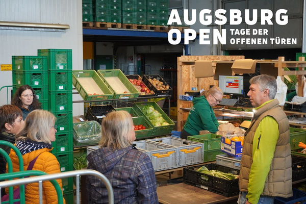 KI generiert: Das Bild zeigt zwei Personen in grüner Arbeitskleidung, die lächelnd eine Kiste mit frischem Obst und Gemüse halten. Im Hintergrund sind weitere Kisten mit Tomaten, Äpfeln und Salat zu sehen, was auf ein Lager oder eine Verkaufsstelle für frische Lebensmittel hinweist.