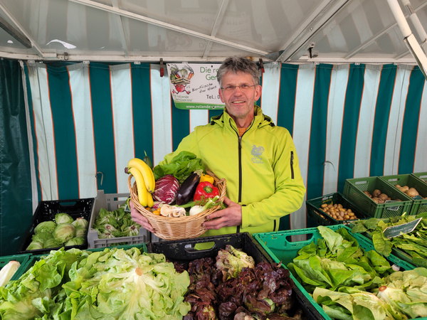 KI generiert: Ein Mann hält einen Korb mit Gemüse auf einem Marktstand. Im Hintergrund sind grüne Kisten mit Gemüse.
