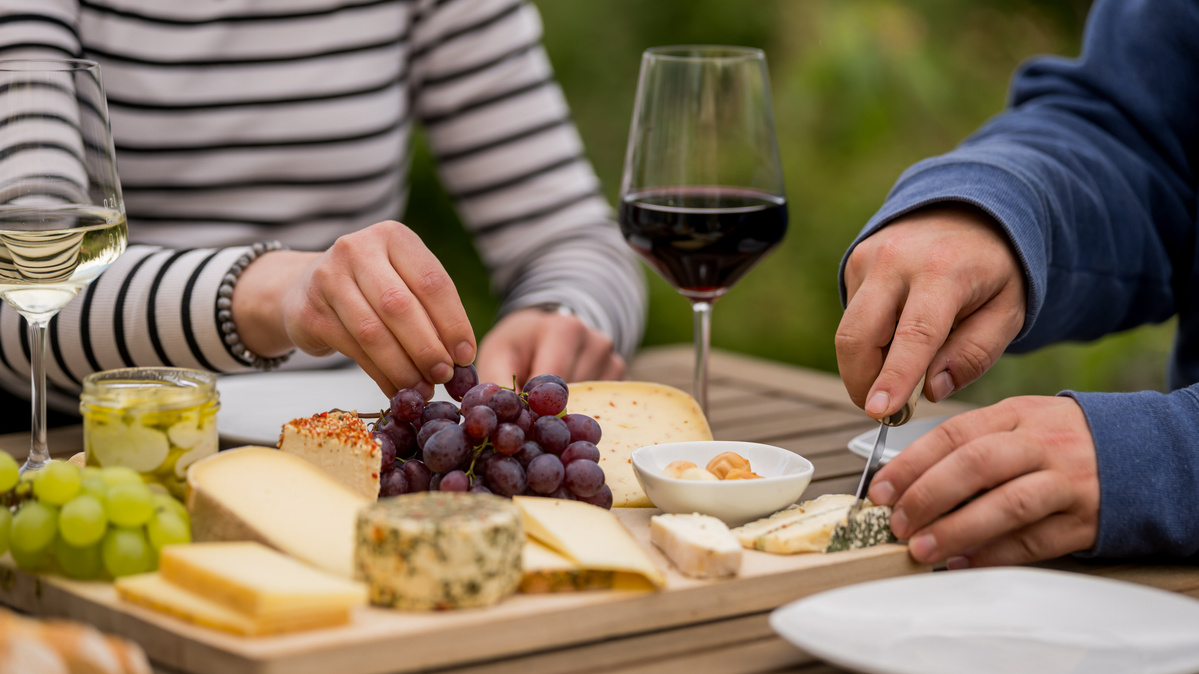 KI generiert: Das Bild zeigt ein hölzernes Brett mit einer Auswahl an verschiedenen Käsesorten, Trauben, Nüssen und Baguettescheiben. Daneben stehen zwei Weingläser, eines davon mit Rotwein gefüllt.