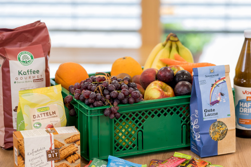 KI generiert: Eine Kiste mit Obst, Kaffee, Snacks und Getränken. Text enthält: "Datteln", "Kaffee Gourmet", "Gute Laune", "HAFER DRINK".