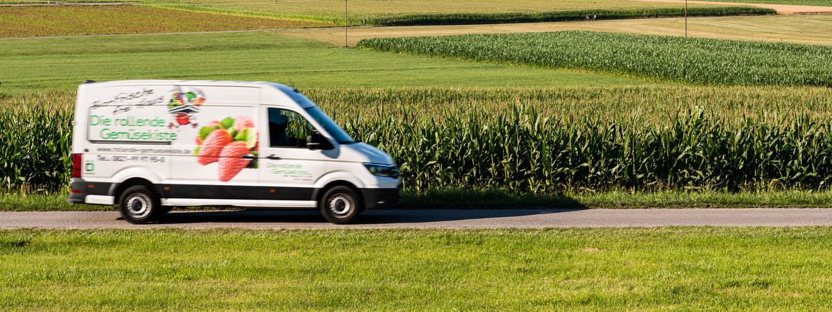 KI generiert: Ein Lieferwagen fährt an einem Feld vorbei. Text: "Die rollende Gemüsekiste".
