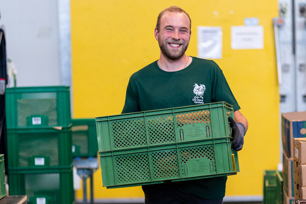 Das Bild zeigt einen Mann mit kurzen, dunklen Haaren und einem freundlichen Lächeln. Er trägt zwei Ökokisten in der Hand.  