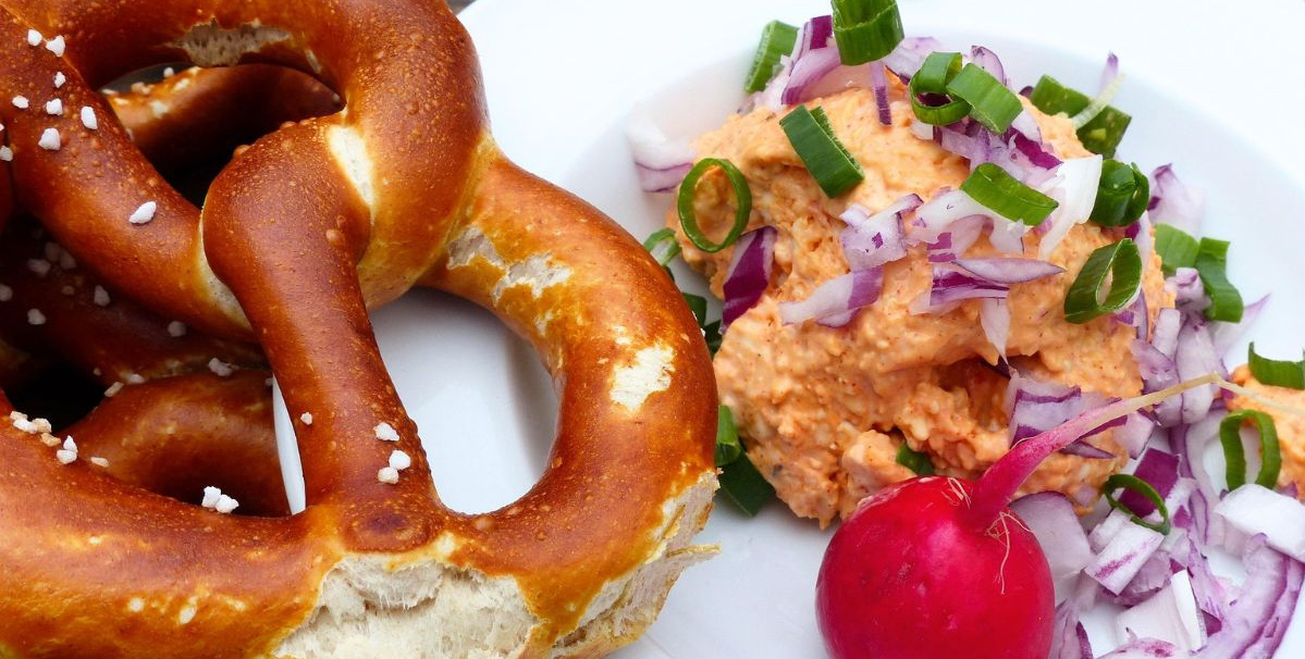 KI generiert: Brezel mit Obazda, Radieschen, roten Zwiebeln und Frühlingszwiebeln auf einem Teller.