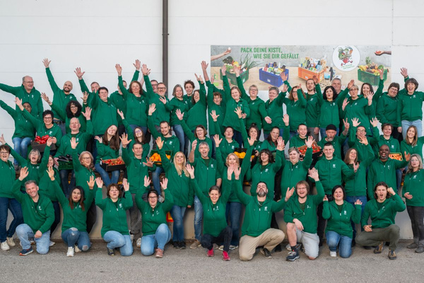 KI generiert: Das Bild zeigt zwei Personen in grüner Arbeitskleidung, die lächelnd eine Kiste mit frischem Obst und Gemüse halten. Im Hintergrund sind weitere Kisten mit Tomaten, Äpfeln und Salat zu sehen, was auf ein Lager oder eine Verkaufsstelle für frische Lebensmittel hinweist.