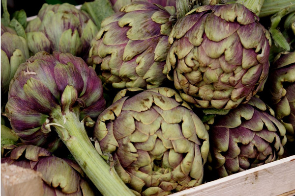 KI generiert: Das Bild zeigt zwei Personen in grüner Arbeitskleidung, die lächelnd eine Kiste mit frischem Obst und Gemüse halten. Im Hintergrund sind weitere Kisten mit Tomaten, Äpfeln und Salat zu sehen, was auf ein Lager oder eine Verkaufsstelle für frische Lebensmittel hinweist.