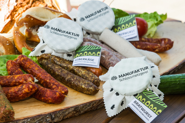 KI generiert: Verschiedene Wurstsorten mit Etiketten "MANUFAKTUR Schlossmetzgerei", "LEBERKÄSE", "BRATWURST" auf einem Holzbrett.