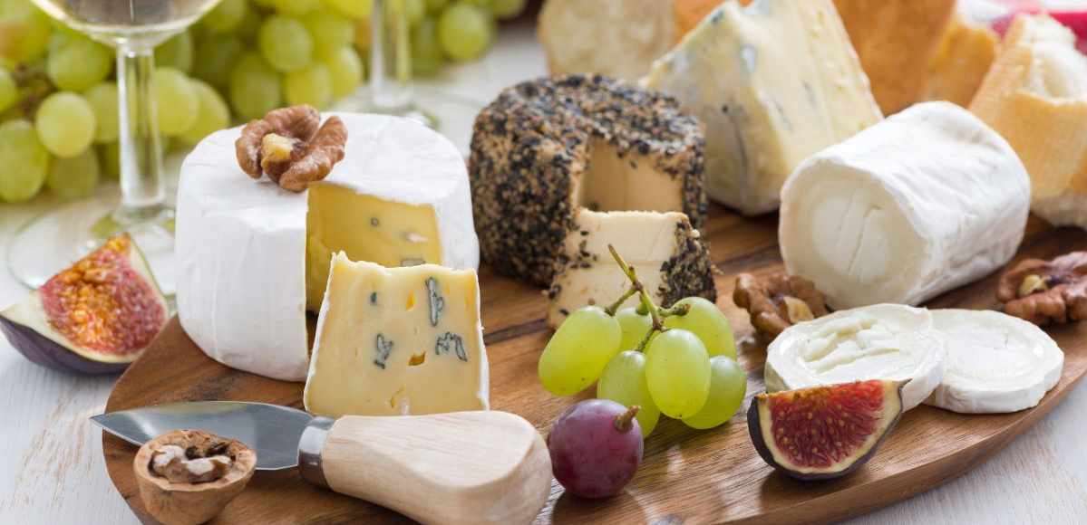 KI generiert: Das Bild zeigt ein hölzernes Brett mit einer Auswahl an verschiedenen Käsesorten, Trauben, Nüssen und Baguettescheiben. Daneben stehen zwei Weingläser, eines davon mit Rotwein gefüllt.