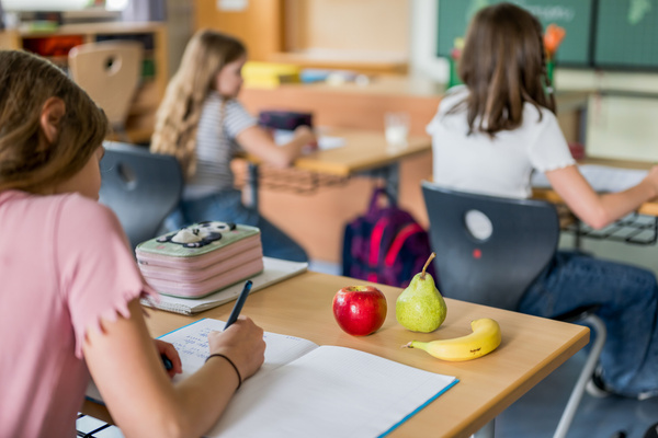 KI generiert: Das Bild zeigt zwei Personen in grüner Arbeitskleidung, die lächelnd eine Kiste mit frischem Obst und Gemüse halten. Im Hintergrund sind weitere Kisten mit Tomaten, Äpfeln und Salat zu sehen, was auf ein Lager oder eine Verkaufsstelle für frische Lebensmittel hinweist.