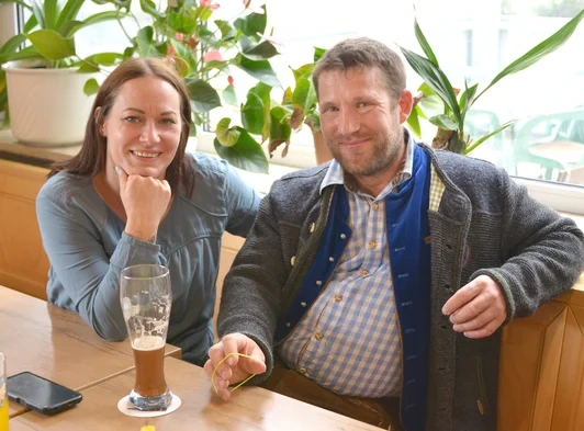 KI generiert: Ein Mann und eine Frau sitzen lächelnd an einem Tisch mit Getränken vor Pflanzen.