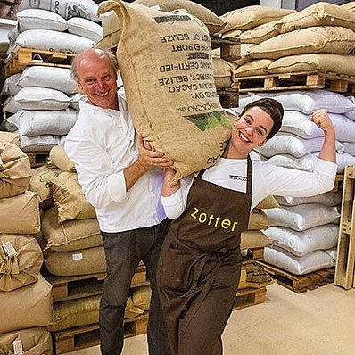 KI generiert: Zwei Personen heben fröhlich einen großen Sack in einer Lagerhalle voller Säcke. Text: "zotter" auf Schürze.