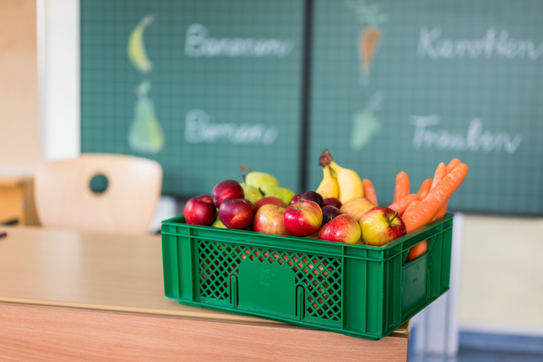 KI generiert: Eine Kiste mit Obst und Gemüse steht auf einem Tisch; Tafel im Hintergrund mit den Wörtern: Bananen, Karotten.