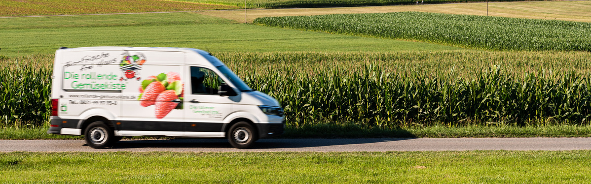 KI generiert: Ein Lieferwagen mit Obstmotiv fährt an einem Feld vorbei. Text: "Die rollende Gemüsekiste".
