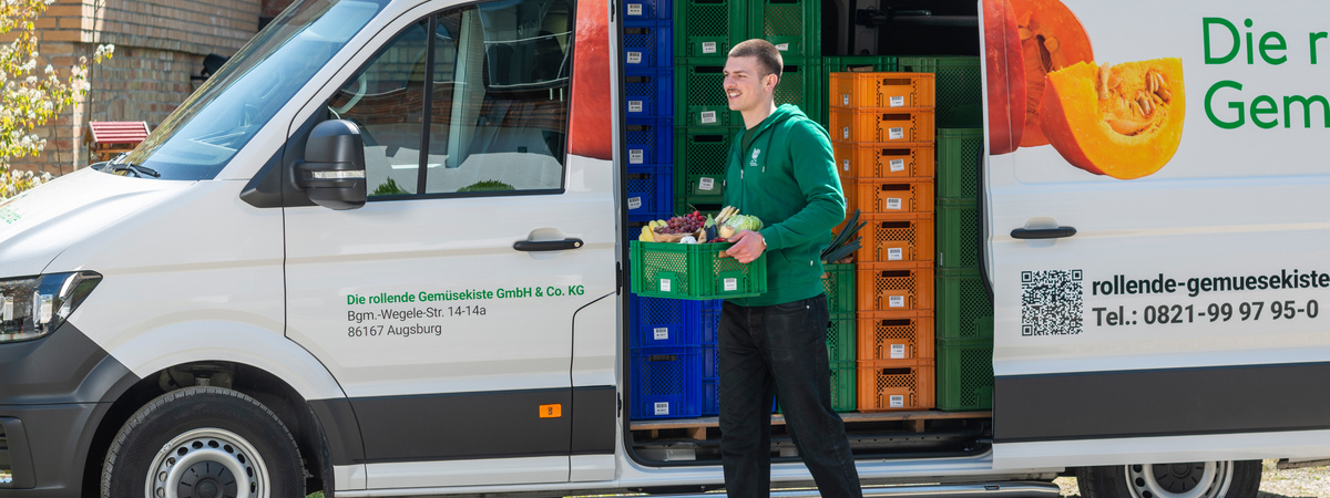 KI-generierte Beschreibung: Ein Mann steht vor einem Ökokisten-Lieferwagen und hält eine Kiste mit Gemüse. Text: "Die rollende Gemüsekiste" und Kontaktdaten.
