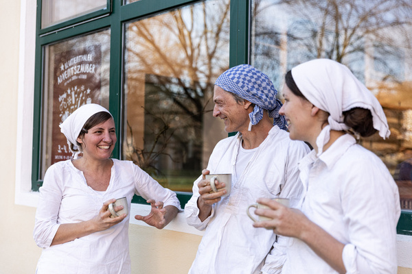 KI generiert: Drei Personen in Weiß mit Kopftüchern trinken Kaffee und unterhalten sich. Text im Fenster: "Allgäu-Adventszauber".