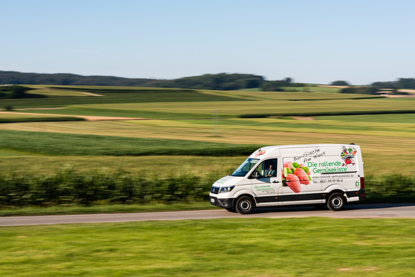 KI generiert: Das Bild zeigt zwei Personen in grüner Arbeitskleidung, die lächelnd eine Kiste mit frischem Obst und Gemüse halten. Im Hintergrund sind weitere Kisten mit Tomaten, Äpfeln und Salat zu sehen, was auf ein Lager oder eine Verkaufsstelle für frische Lebensmittel hinweist.