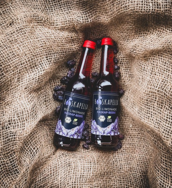 KI generiert: Zwei Flaschen Bio-Limonade „Hoskapella“ auf Jutesack. Text: „BIO-LIMONADE HASKAP-BEERE 0,33l 100% VEGAN“.