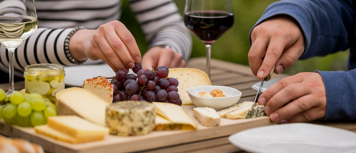 KI generiert: Eine Käseplatte mit Trauben, Nüssen und Wein auf einem Tisch, die von Personen genossen wird.