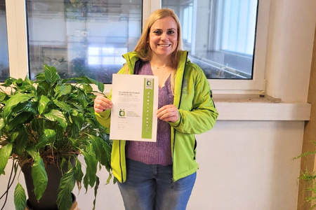 ine Person hält ein Zertifikat neben einer Pflanze. Text: "Zertifikat 2025 Ökokiste".