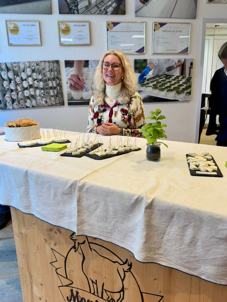 KI generiert: Eine Frau steht hinter einem Tisch mit Käseproben. Im Hintergrund hängen Auszeichnungen an der Wand.