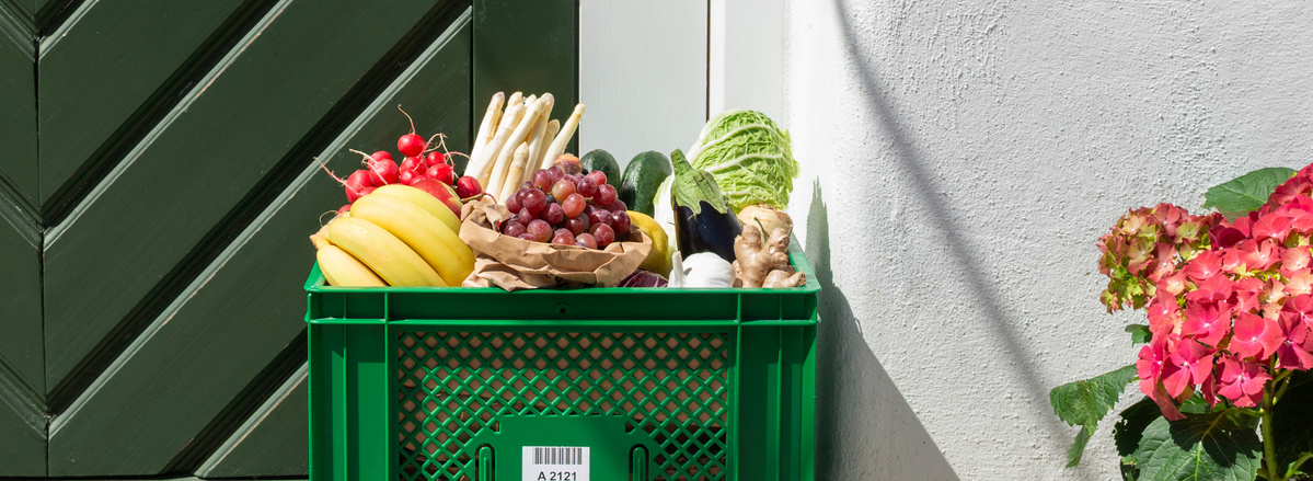 KI generiert: Grüne Kiste mit frischem Obst und Gemüse vor einer Tür, neben blühenden Hortensien.