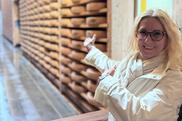 KI generiert: Das Bild zeigt zwei Personen in grüner Arbeitskleidung, die lächelnd eine Kiste mit frischem Obst und Gemüse halten. Im Hintergrund sind weitere Kisten mit Tomaten, Äpfeln und Salat zu sehen, was auf ein Lager oder eine Verkaufsstelle für frische Lebensmittel hinweist.