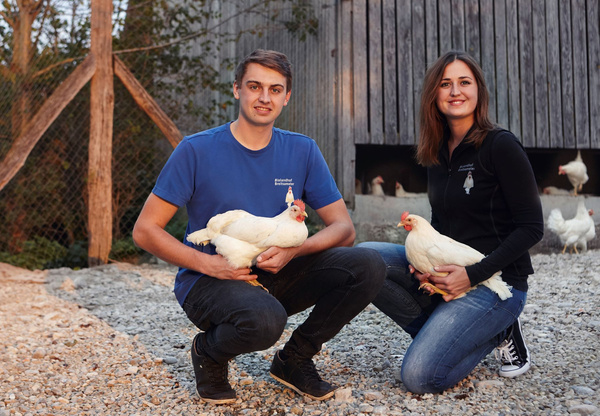 KI generiert: Zwei Personen halten Hühner, umgeben von weiteren Hühnern vor einem Stall.