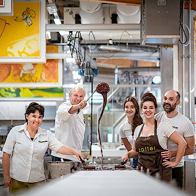 KI generiert: Fünf Personen in einer Schokoladenfabrik lächeln, während eine Person Schokolade gießt.