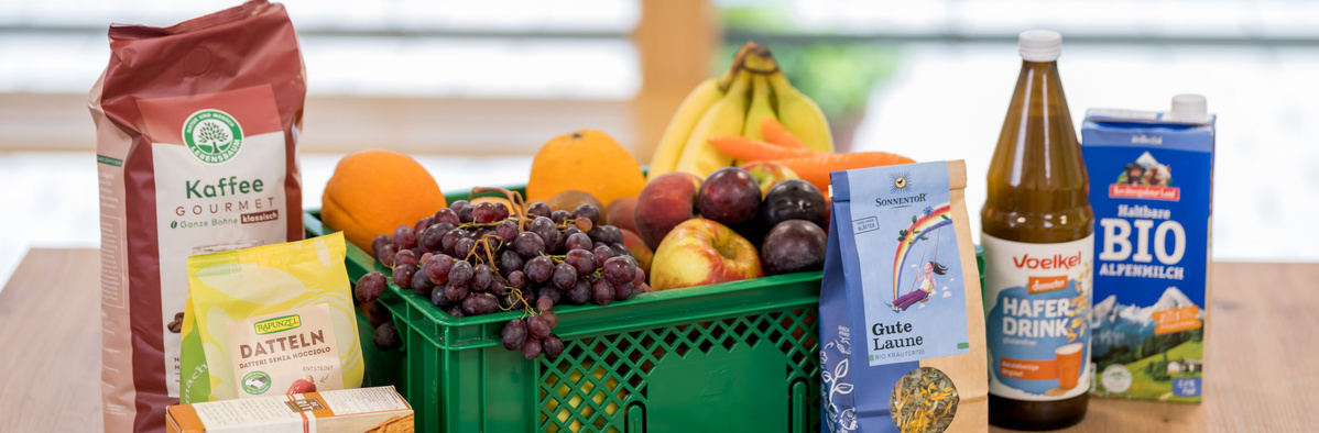 KI generiert: Eine Gemüsekiste mit Obst, Kaffee, Datteln und Getränken. Texte: "Kaffee", "Dateln", "Hafertdrink", "BIO".