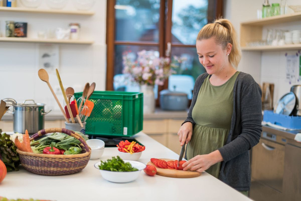 KI generiert: Das Bild zeigt zwei Personen in grüner Arbeitskleidung, die lächelnd eine Kiste mit frischem Obst und Gemüse halten. Im Hintergrund sind weitere Kisten mit Tomaten, Äpfeln und Salat zu sehen, was auf ein Lager oder eine Verkaufsstelle für frische Lebensmittel hinweist.