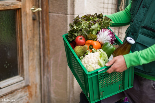 KI generiert: Das Bild zeigt zwei Personen in grüner Arbeitskleidung, die lächelnd eine Kiste mit frischem Obst und Gemüse halten. Im Hintergrund sind weitere Kisten mit Tomaten, Äpfeln und Salat zu sehen, was auf ein Lager oder eine Verkaufsstelle für frische Lebensmittel hinweist.