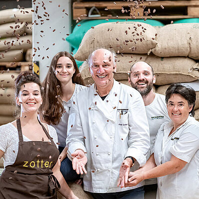 KI generiert: Menschen mit Kakaobohnen vor Kakaosäcken; Text auf Schürze: "zotter".