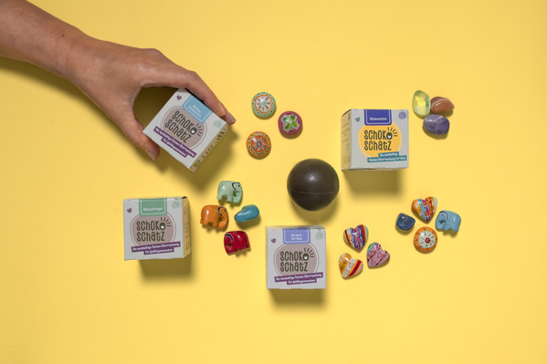 KI generiert: Farbige Schokoladenverpackungen und bunte Accessoires auf gelbem Hintergrund. Text: "Schoko Schatz".