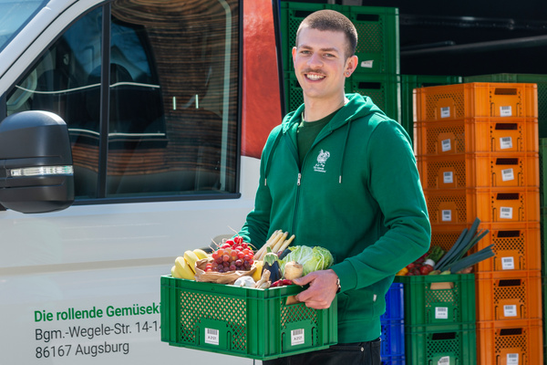 KI generiert: Eine lächelnde Tomate trägt eine Kiste mit Lebensmitteln. Text: "Die rollende Gemüsekiste".