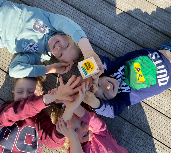 KI generiert: Kinder liegen auf dem Boden, lächeln und strecken ihre Hände in die Mitte aus. Text: "SCHOOLOGY SUPERSTAR".