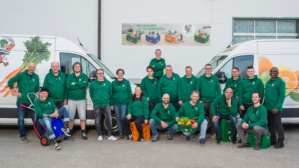 KI generiert: Eine Gruppe von Menschen posiert in grünen Jacken vor zwei Lieferwagen. Text: "PACK DEINE KISTE, WIE SIE DIR GEFÄLLT."