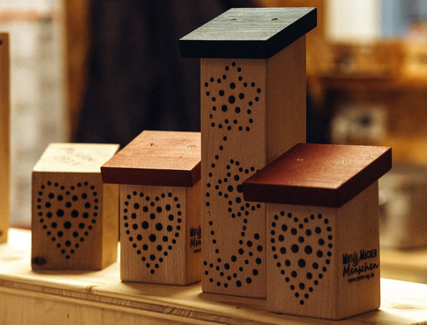 KI generiert: Holzinsektenhotels mit dekorativen Löchern, einige haben rote und schwarze Dächer. Text: "Mönchmühle".