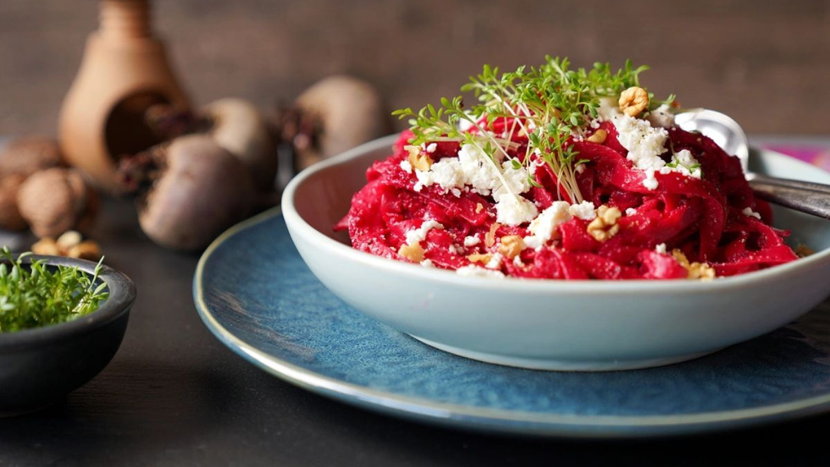 KI generiert: Das Bild zeigt eine Schüssel mit einem bunten Salat aus Rote-Bete-Scheiben, Karottenstreifen, Feldsalat, gehackten Nüssen und einem Klecks Sahne oder Joghurt in der Mitte. Der Salat liegt auf einem Holztisch.