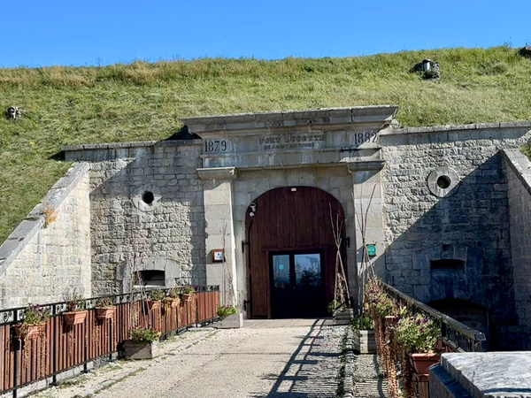 KI generiert: Historischer Steineingang eines Gebäudes, umgeben von Rasen. Text: "1879", "1882", "FORT ECOSSAIS".