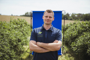 KI generiert: Ein Mann steht mit verschränkten Armen vor einem blauen Hintergrund in einem Feld. Sein Shirt zeigt "Pechler BIOHOF".